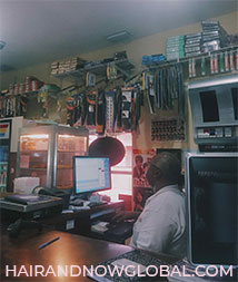 Inside the corner store in Grand Cayman, male shop keeper sitting at the counter surrounded by basic groceries and a variety of wigs and hair extensions hanging from ceiling